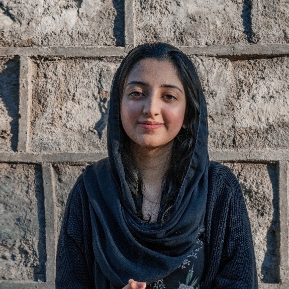 Portrait of a young person dressed in black with a headscarf, looking straight into the camera with a confident smile.