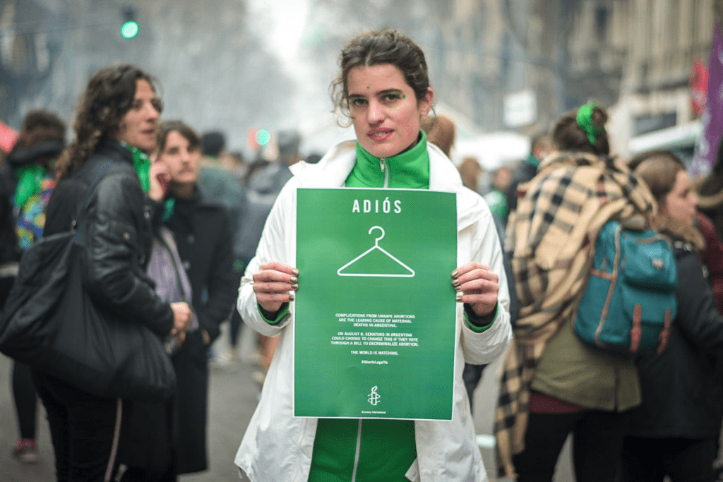 someone at a protest holding the image of the Amnesty advertisement on the new york times. At the top of the ad it reads Adios, with an icon of a coat hanger. 