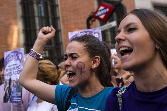 two women are in the focus of a photo taken at a protest. The one on the left has a purple Venus symbol painted on her cheek and has one of her hands in the air in a fist. 