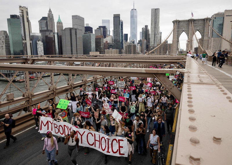 A crowd of protesters proceeds over the Brooklyn Bridge with the New York skyline in the background. They carry a large banner with the words 'my body my choice'. 