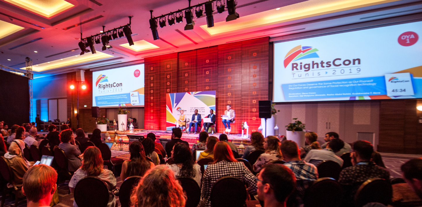 A conference full of people facing a screen which says 'RightsCon'