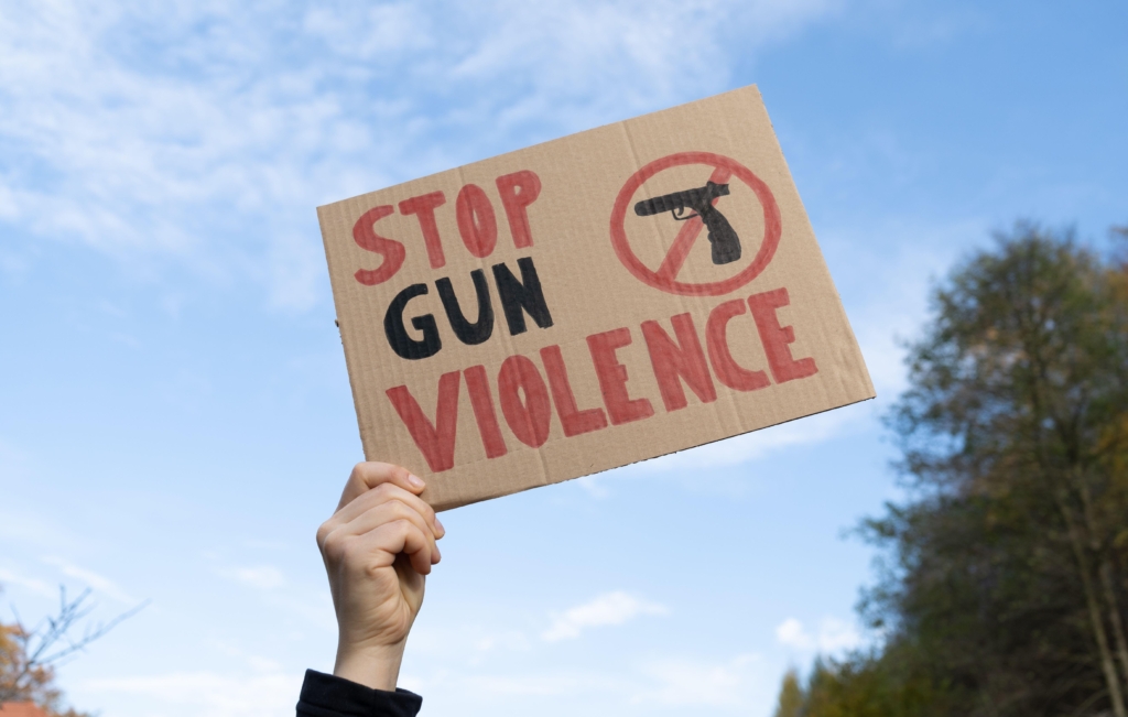 a hand holding a cardboard sign that reads "Stop Gun Violence" and has an icon of a gun which is crossed out. 