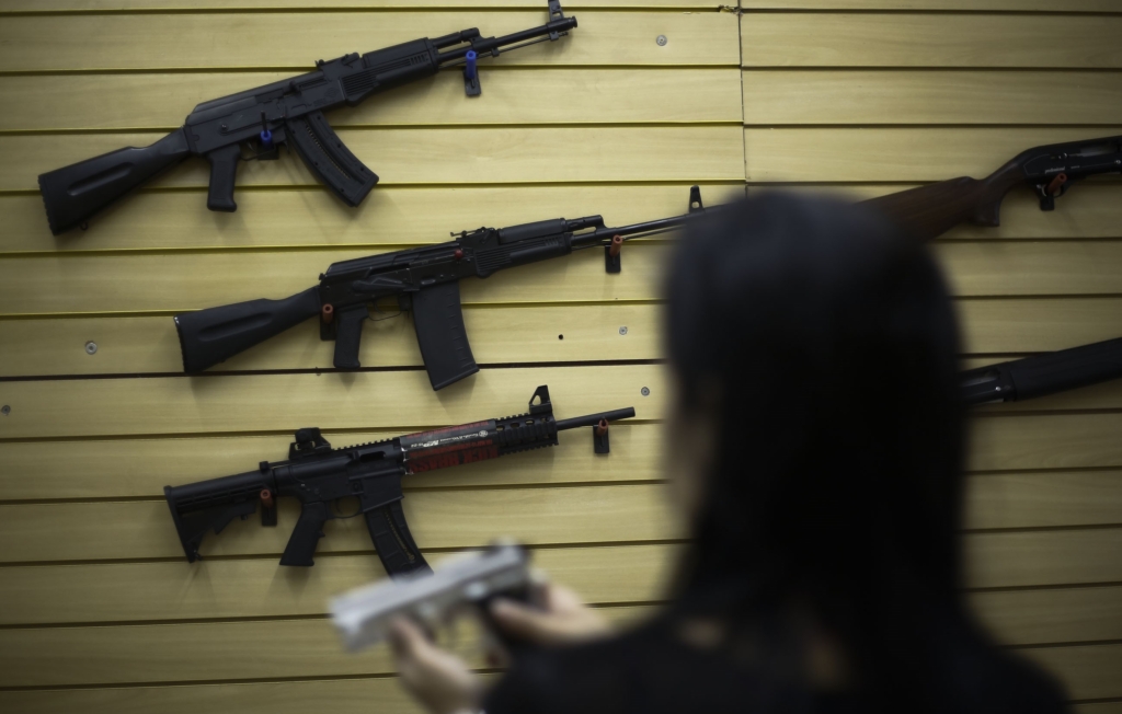a collection of rifles are mounted on a wall in a gun shop in Brazil. There is a woman in the forefront of the picture who is holding a pistol.  