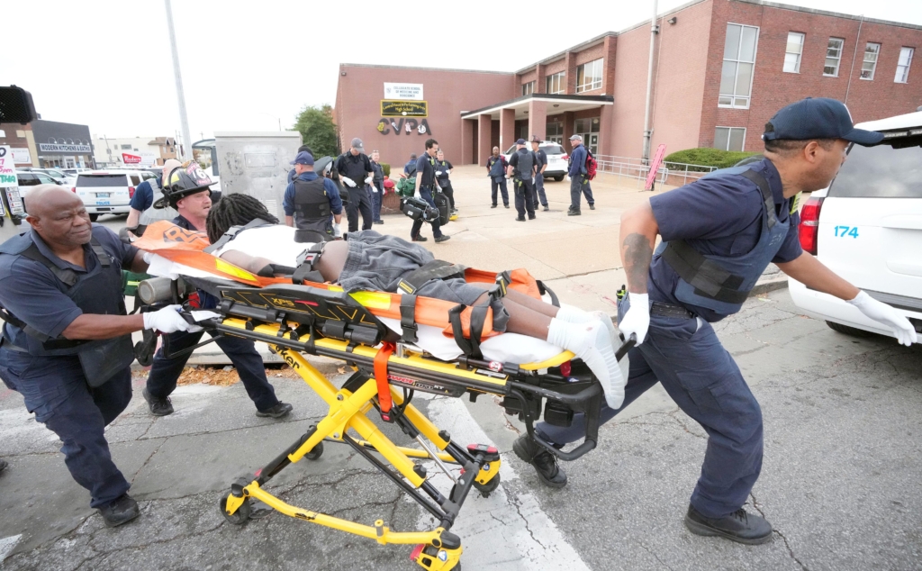 a shooting victim lying face down on a stretcher and is being escorted by two firefighters. Both of the firefighters are wearing navy bullet proof vests