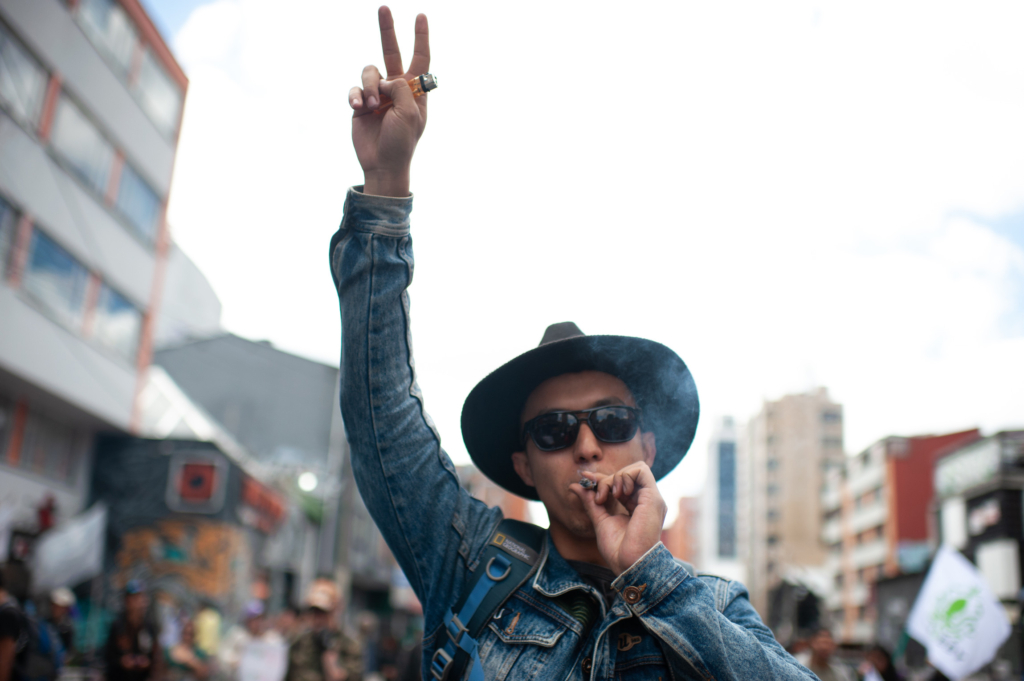 someone is participating in a march and is holding the peace sign in the air. They are wearing a wide brimmed hat, sunglasses and are smoking. 