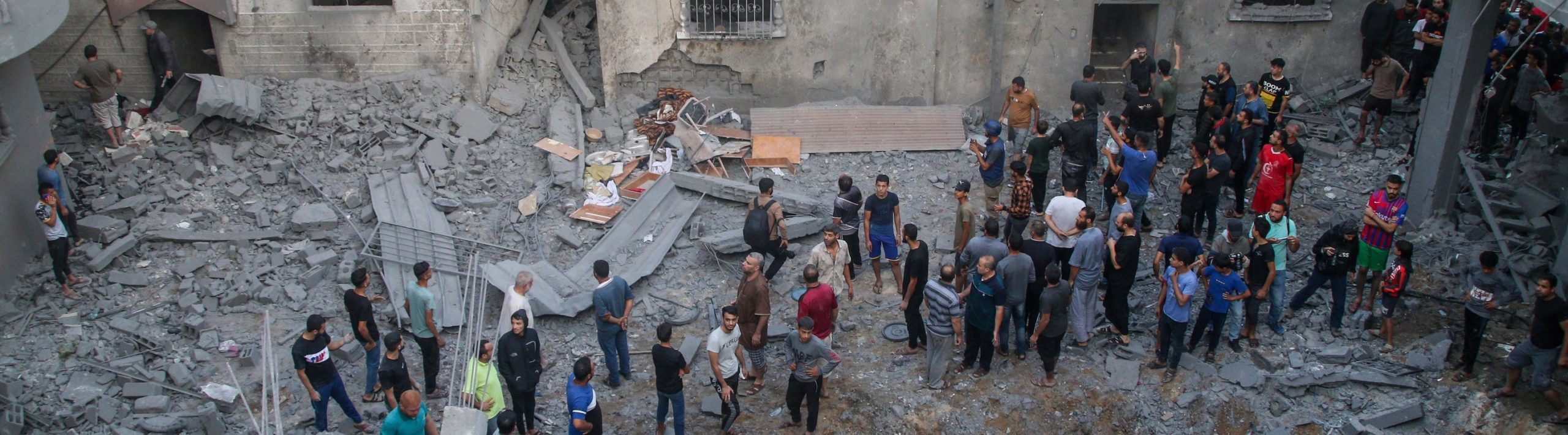 A crowd of people standing amongst rubble, and bombed building.