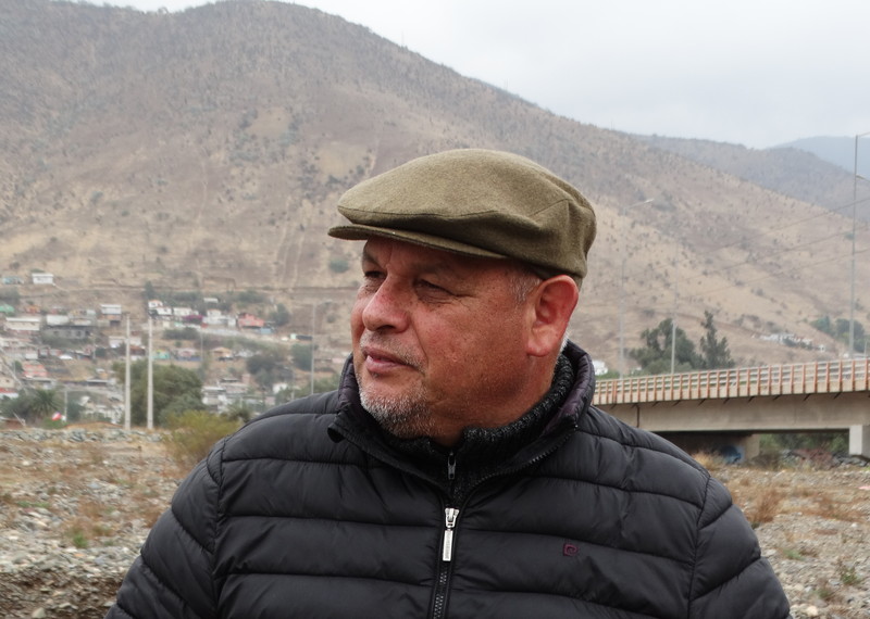 A man wearing a coat and hat stands looking to the side, with a bridge and a mountain in the background.