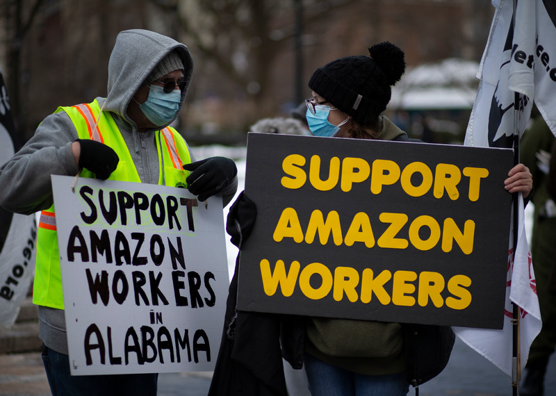 Des manifestant·e·s masqué·e·s portent des pancartes qui affichent « Soutenez les travailleurs d’Amazon ».