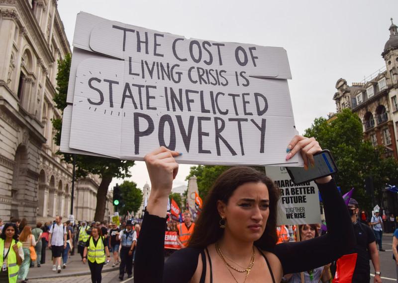 Une personne au milieu d’une manifestation brandit une pancarte sur laquelle on lit en anglais « la crise du coût de la vie, c’est la pauvreté infligée par l’État »