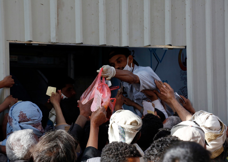 A crowd of outstretched hands reach towards a plastic bag of food offered by a uniformed worker behind a hatch.