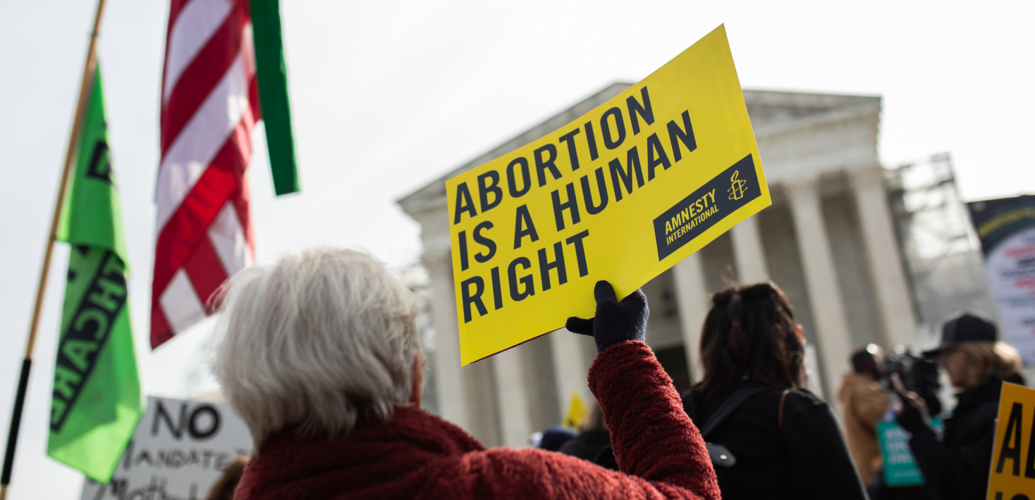 A person with short grey hair, holds up a yellow sign that says abortion is a human right outside, while a US flag flies high to her left.