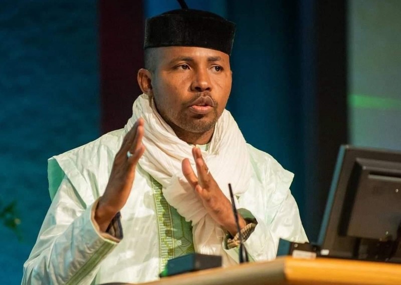 Daouda Diallo stands at a podium and talks into a microphone. He is wearing a black hat and has a white scarf tied stylishly round his neck.