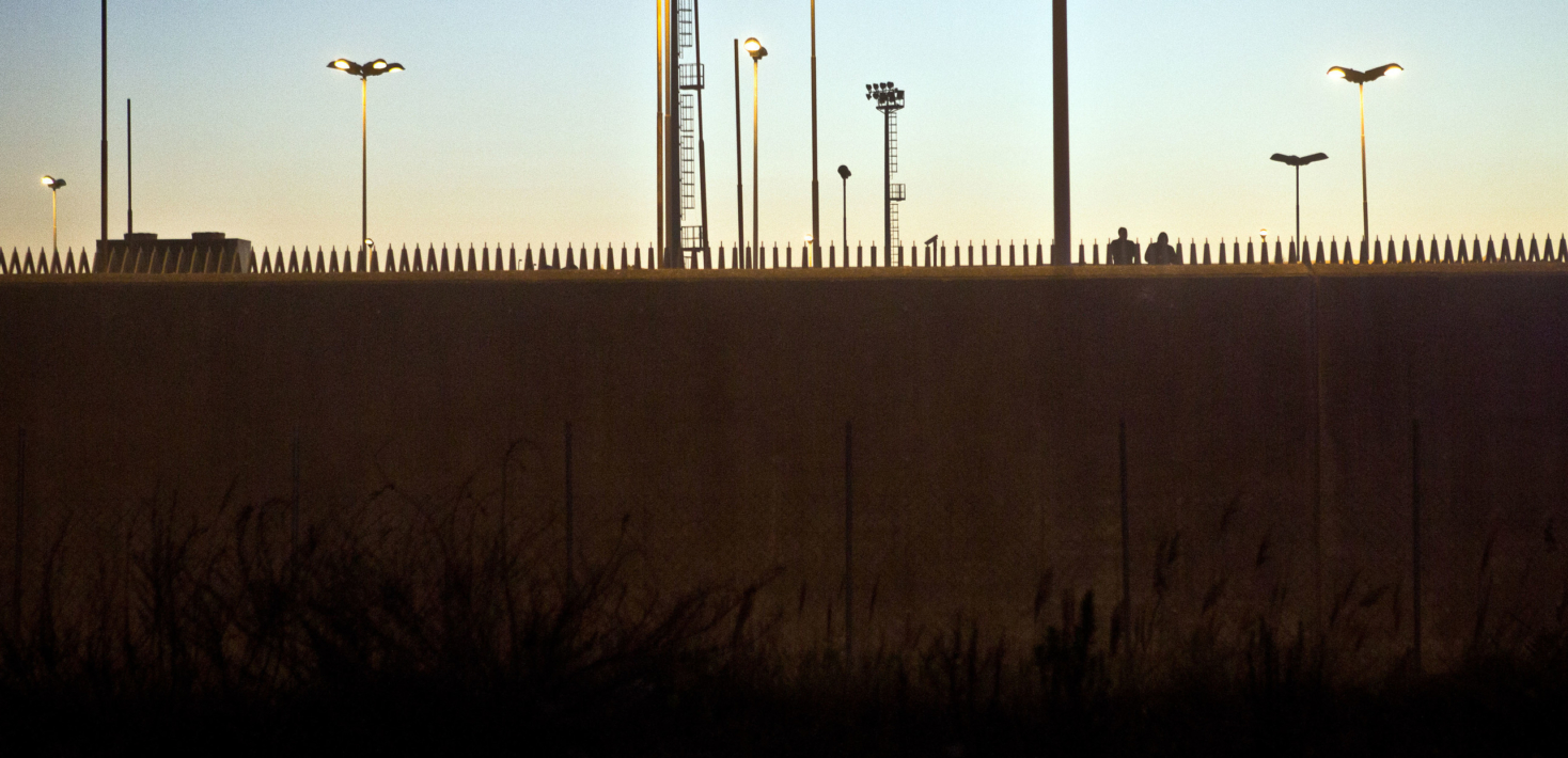 Barbed wried walls surrounding a detention centre with two people in the distance looking over
