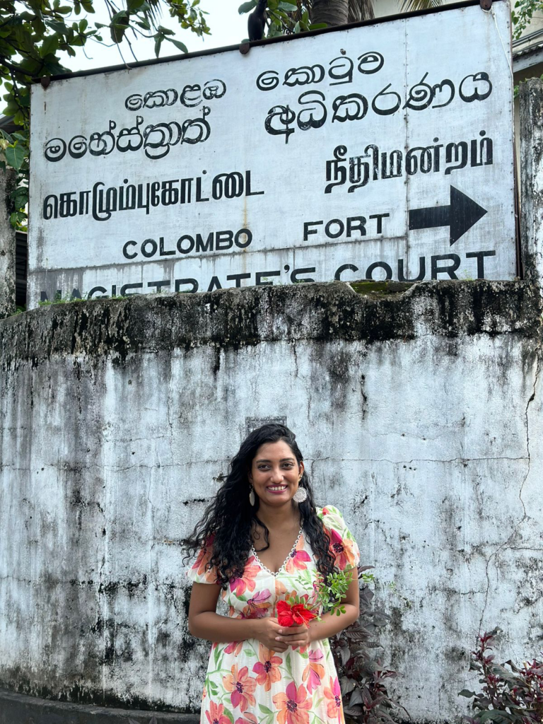 Sri Lankan comedian Nathasha Edirisooriya wears a flow ery dress andstands in front of a tarnished white wall, holding a bunch of red flowers.