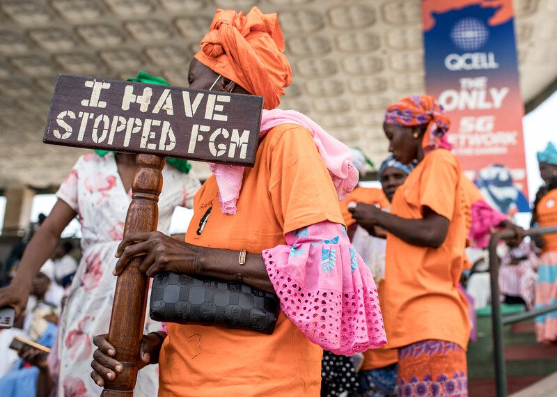 Una mujer de Gambia, con atuendo naranja y rosa, sostiene un cartel en el que se lee “I have stopped FGM” (He puesto fin a la mutilación genital femenina).