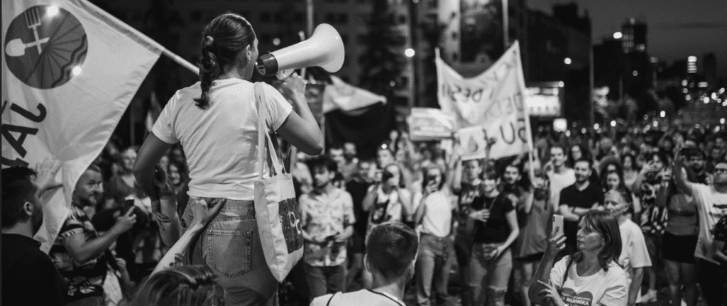 Фотография демонстрации. Один из протестующих говорит в мегафон. Толпа протестующих наблюдает за тем, как этот человек говорит.