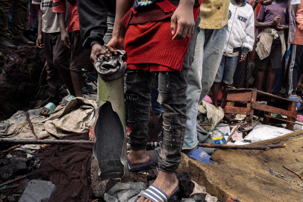 A boy standing on a mattress amid broken wooden sticks and torn clothing, holds a ripped and crumpled metal tube from a rocket, about as long as one of his legs, surrounded by a group of onlookers.