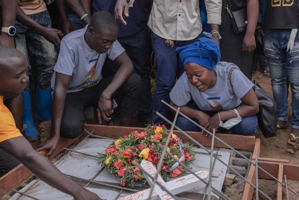 A woman in a blue headwrap, kneeling on the ground, cries over a grave with a cross and a bouquet of flowers, as other mourners surround her.