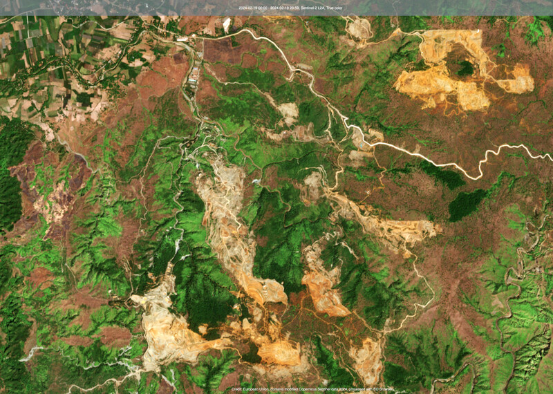 Satellite image showing large green areas of forest and a settlement with houses and farmland in the top left hand corner. Throughout the image are big areas of land where trees have been removed to make way for nickel mines, which are yellow and gold in colour from this distance