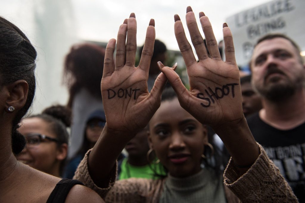 someone in a crowd holds their hands up. On their hands you can read the words 'Don't Shoot'