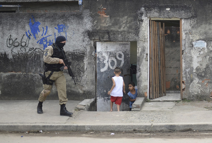 Dos niños se apresuran a entrar en una vivienda ante la presencia de un agente armado que avanza por la calle con un rifle de gran tamaño.