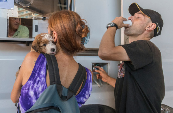 Dos pacientes ante una ventanilla, en la que pueden recoger medicación para el tratamiento contra la drogadicción. La mujer sostiene un cachorro, mientras el hombre bebe un medicamento de un vaso de plástico blanco.